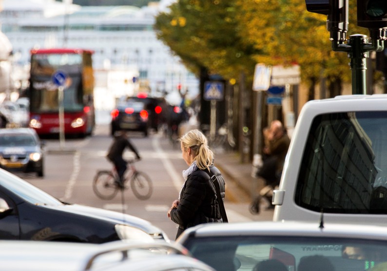 gatuvy med bilar, buss, cykel och en g&aring;ngtrafikant, en kvinna med svart kappa och blont h&aring;r i h&auml;stsvans
